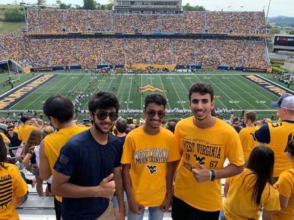 Group of 3 international students wearing gold and blue posing at a WVU football game.